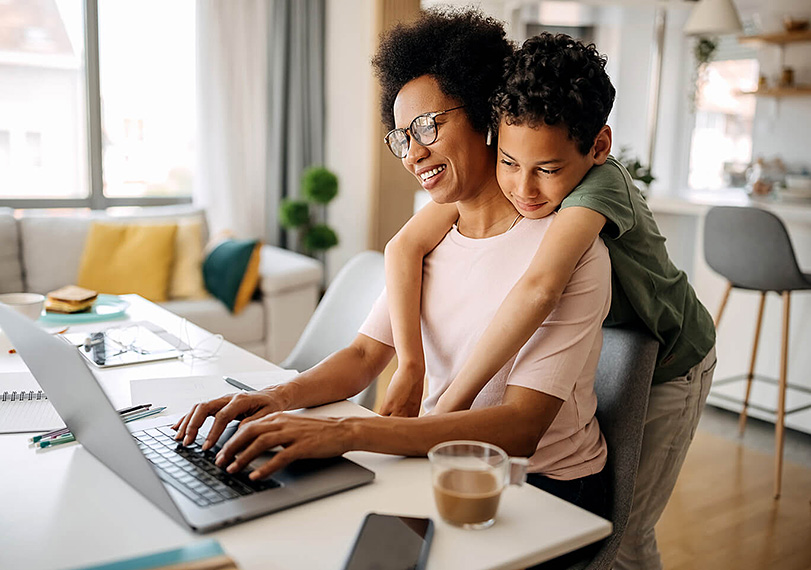 Parent using a laptop at home while a child leans in with a hug from behind