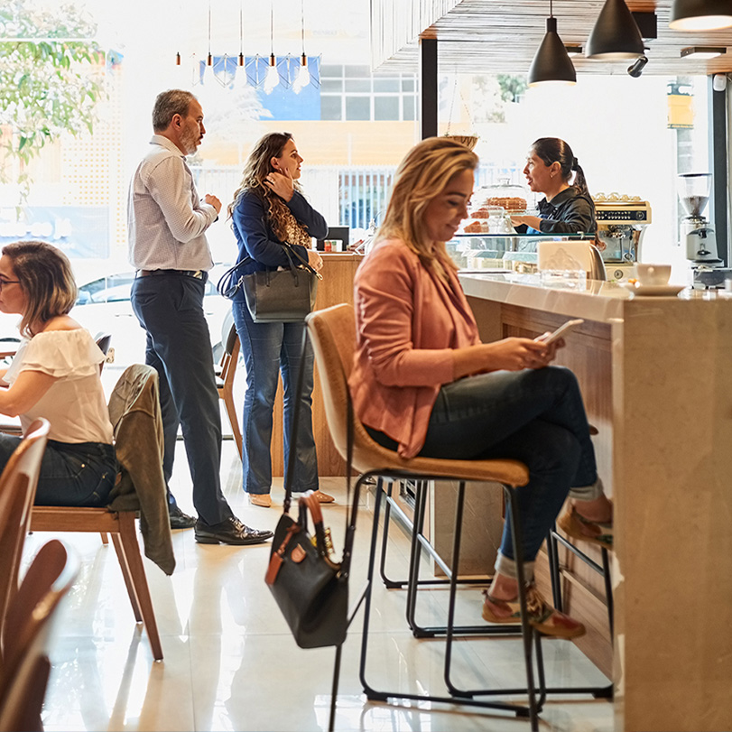 People waiting and sitting in a café, including a woman using her phone at the counter